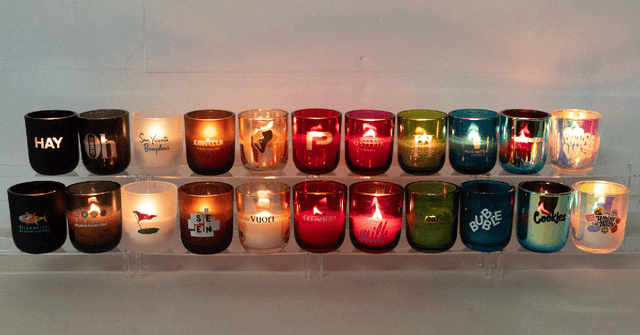 A display of colorful glass candles, some lit and some unlit, arranged in two rows on clear shelves against a plain wall. Each candle has a unique design or text on it.