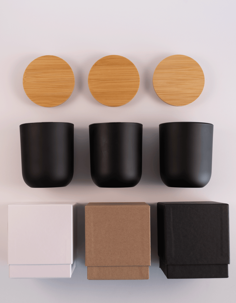 Three black glass tumblers with wooden lids above and three different boxes; white, kraft, and black; on a white surface.