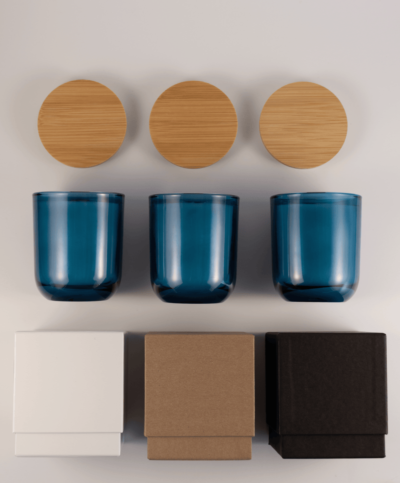 Three blue glass tumblers with wooden lids above and three different boxes; white, kraft, and black; on a white surface.