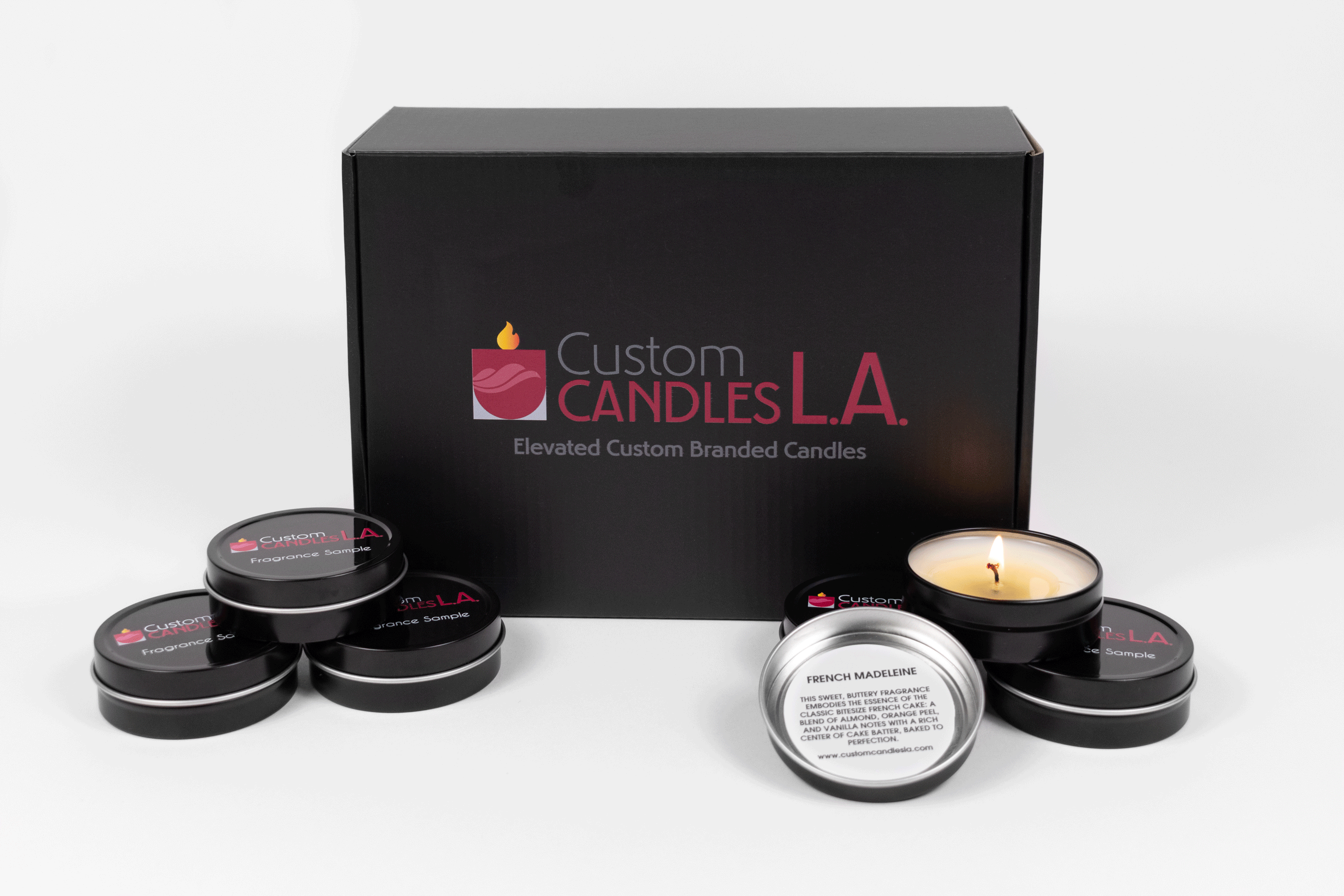 Fragrance samples box from Custom Candles L.A. The black cardboard box is sitting on its side, so the top is facing to the front. Printed on the box is the Custom Candles L.A. Logo in burgundy and grey with "Elevated Custom Branded Candles" printed below the logo. To the left and right of the box are stacks of 3 black fragrance sample tins. The top sample on the right is a lit candle sample with the lid removed that reads "French Madeleine" on the inside of the lid and its description.