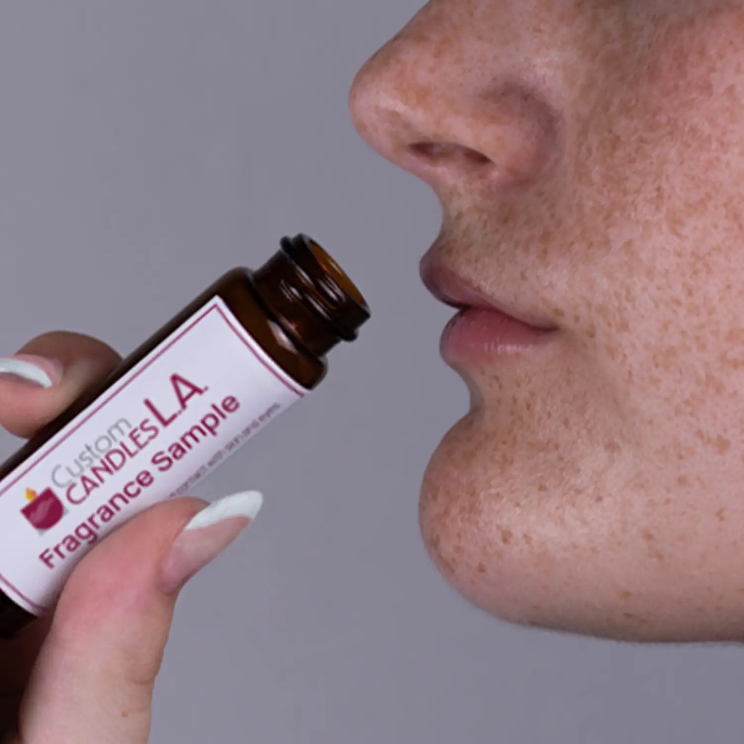 Person smelling a Custom Candles LA fragrance sample bottle close to their nose against a gray background