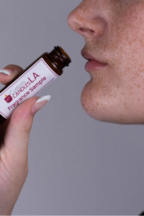 Person holding a Candles LA fragrance sample bottle close to their nose against a gray background