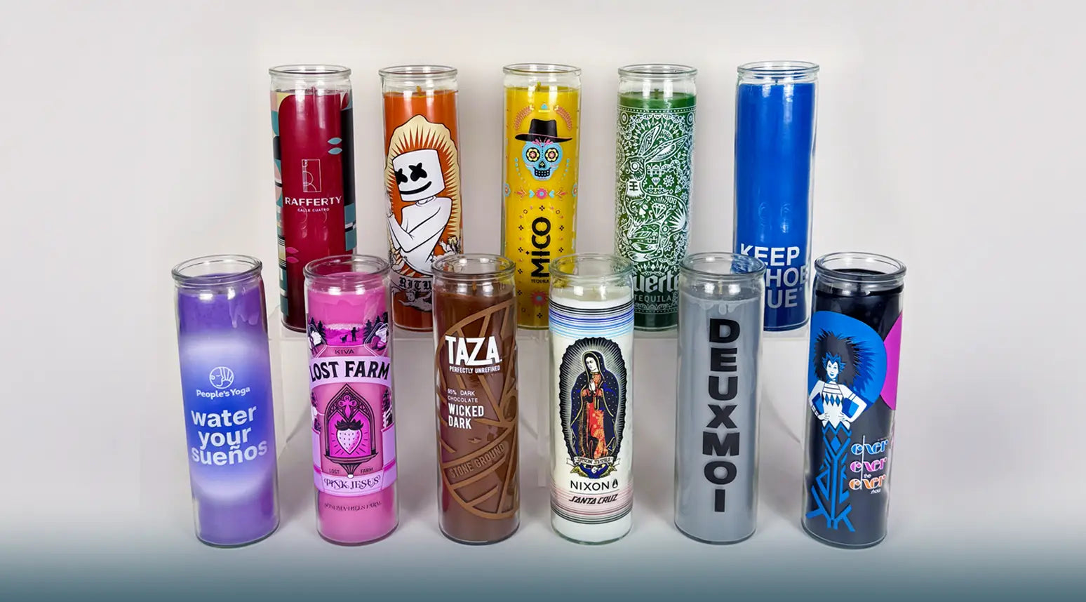 a group shot of 11 prayer candles all with different color waxes and different logos and designs on each one, arranged in two tiers with 6 being in front and 5 being behind on an elevated platform