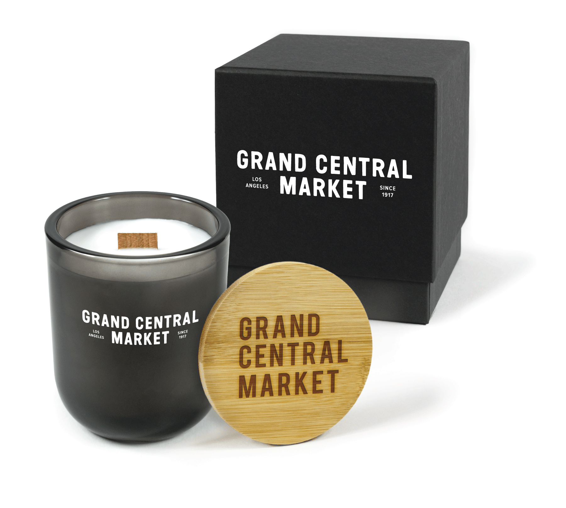 A 12 oz Luxe Custom Candle in the color “Nightfall” which is a smoke gray from Custom Candles LA. The candle features a wood wick and white logo for “Grand Central Market” with “Los Angeles” and “Since 1917" on the sides printed on the front of the glass. Leaning against the side of the candle is a bamboo candle lid engraved with “Grand Central Market”. In the background, there is a black 2-piece box printed with the full “Grand Central Market” logo.