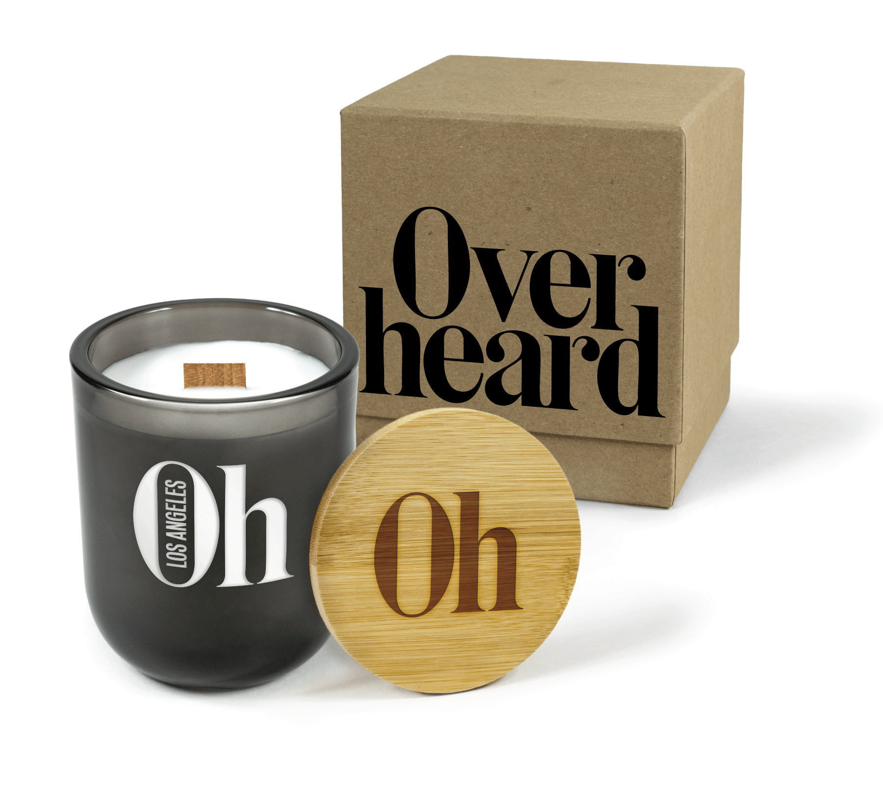 A 12 oz Luxe Custom Candle in the color “Nightfall” which is a smoke gray from Custom Candles LA. The candle features a wood wick and a laser-engraved icon logo for “Overheard Los Angeles” on the front of the glass allowing the white candle wax to show through. Leaning against the side of the candle is a bamboo candle lid engraved with the “Oh” icon logo. In the background, there is a kraft 2-piece box printed with a black “Overheard” logo.