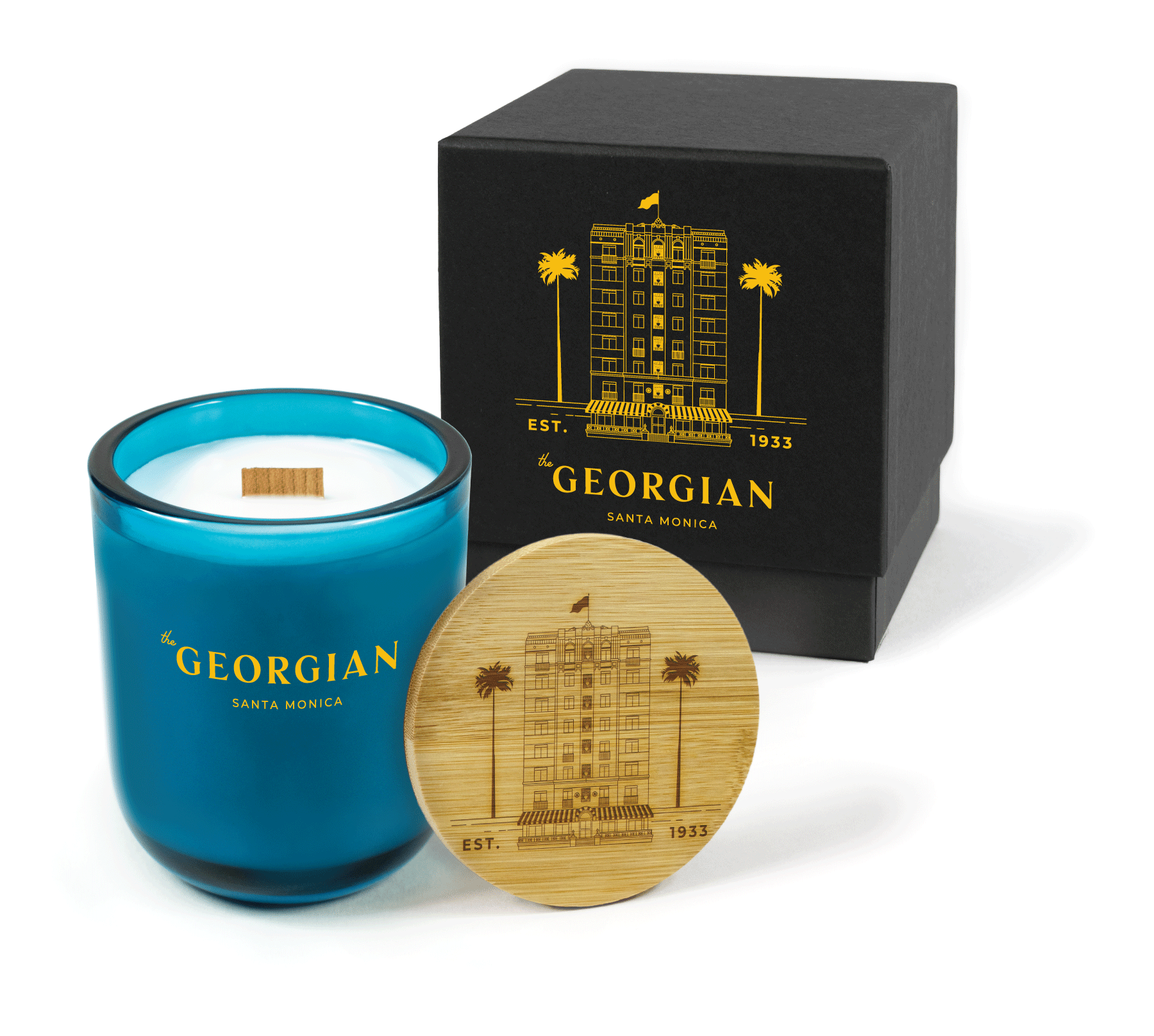 A 12 oz Luxe Custom Candle in the color “Oceania” which is a splashy teal from Custom Candles LA. The candle features a wood wick and 4-color golden yellow logo for “The Georgian Santa Monica” printed on the front of the glass. Leaning against the side of the candle is a bamboo candle lid engraved with and intricate drawing of the building with “Est 1933”. In the background, there is a black 2-piece box printed with a golden yellow version of the intricate building drawing and “The Georgian” logo.
