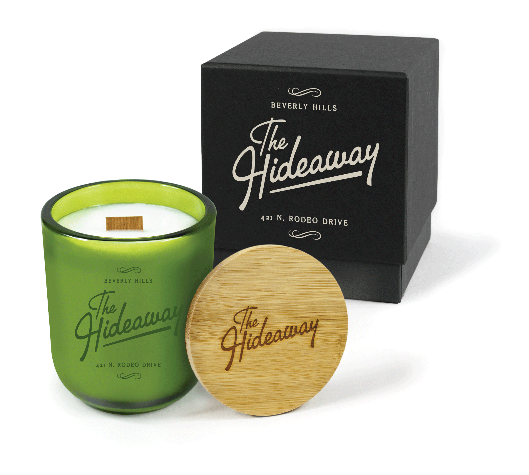 A 12 oz Luxe Custom Candle in the color “Grassland” which is a verdant green from Custom Candles LA. The candle features a wood wick and a green 4-color imprinted logo for “The Hideaway” in Beverly Hills on the front of the glass. Leaning against the side of the candle is a bamboo candle lid etched with “The Hideaway” logo. In the background, there is a black 2-piece box 4-color printed with a cream color logo for “The Hideaway.”