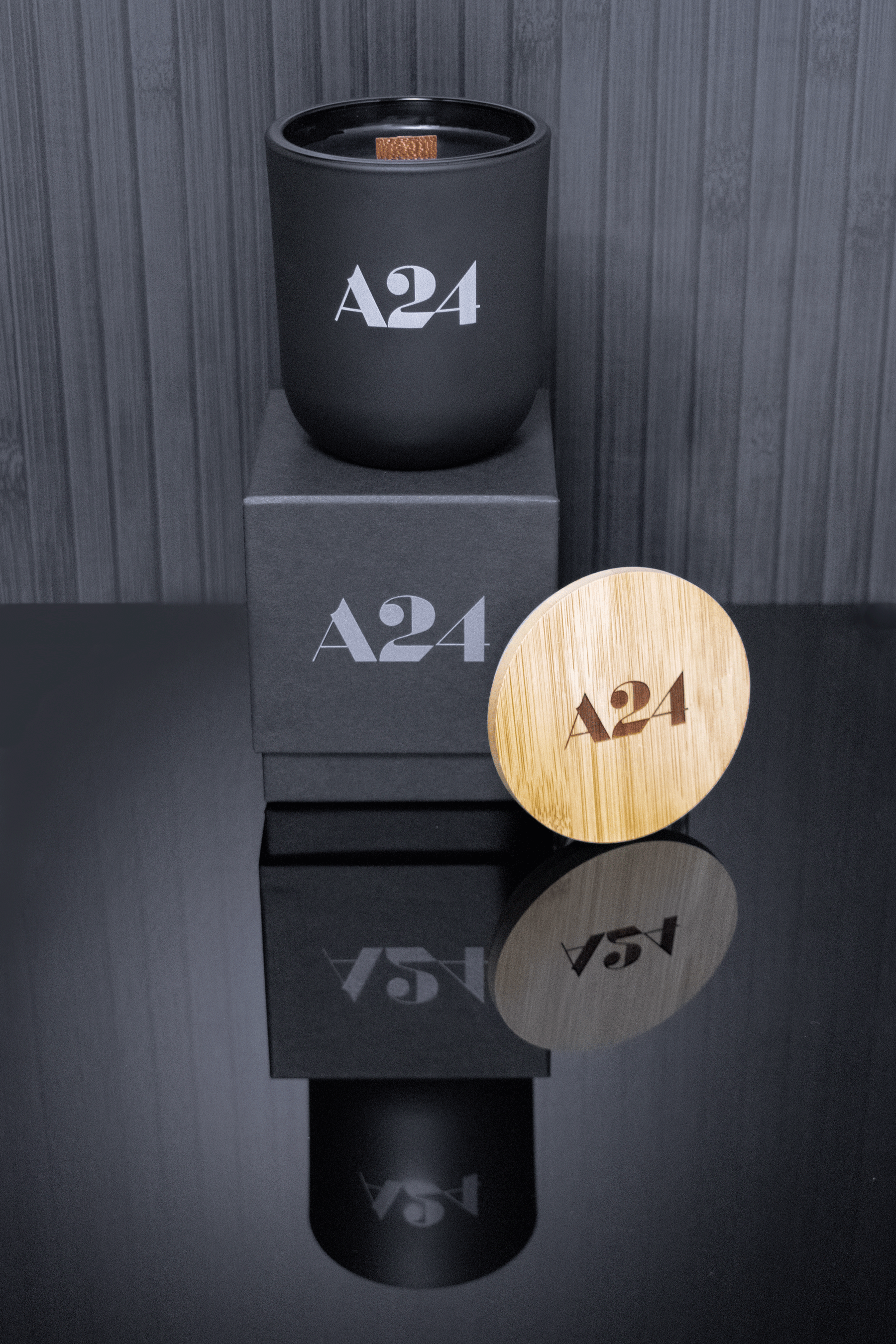 A 12 oz Luxe Candle in "Darkest Night" a jet black color from Custom Candles L.A with black wax and a wooden wick. The candle is printed with a logo that reads "A24" in grey. It sits on a black box that has the same logo printed in grey. Leaning on the right side of the box is a wooden candle lid that is engraved with the same logo. All three items sit on a reflective gray floor, vividly reflecting the candle, box, and lid, and the background are grey wooden vertical slats.