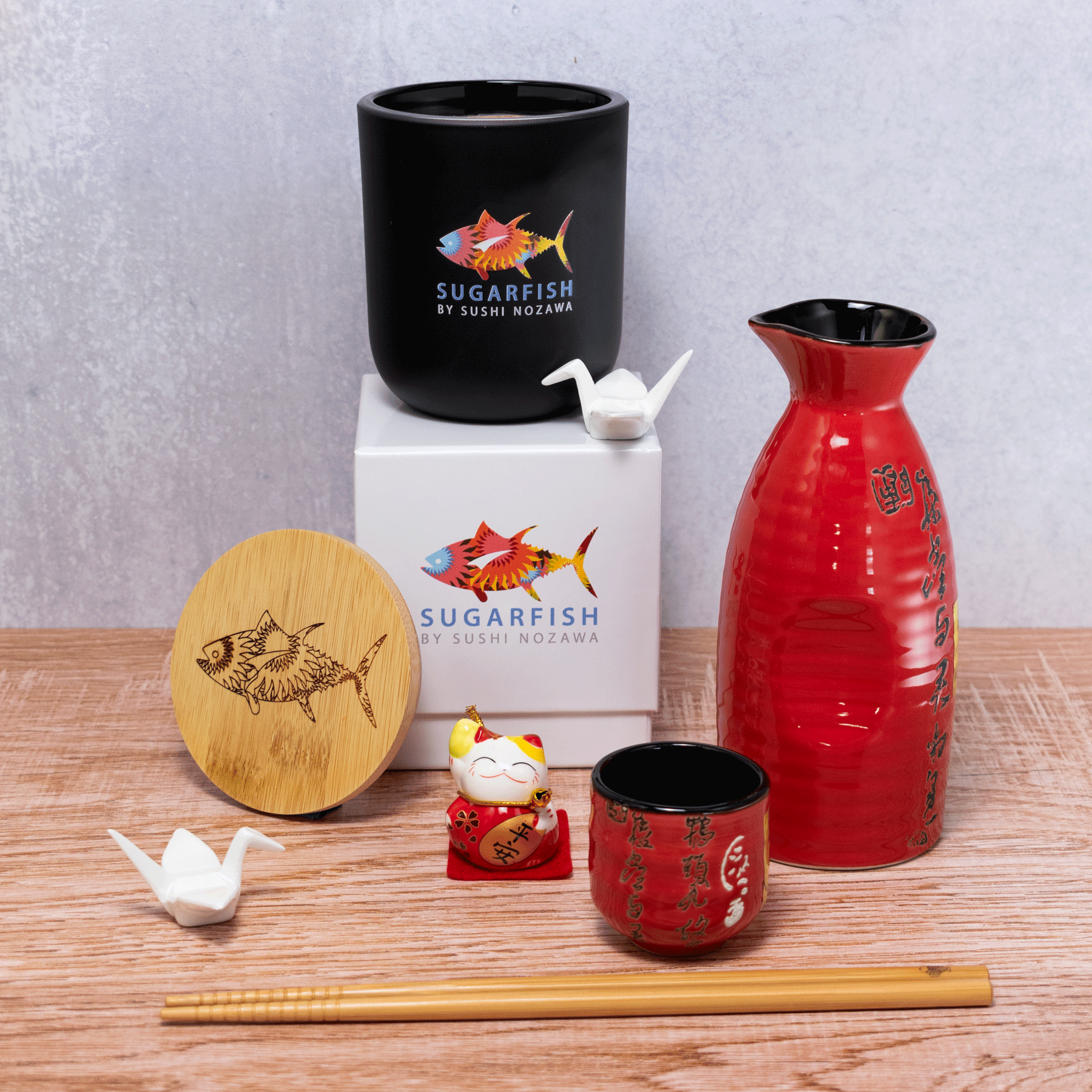 A 12 oz Luxe Candle in "Darkest Night" a jet black color from Custom Candles L.A. The candle is printed with a colorful logo of "sugarfish by sushi nozawa". It sits next to a white crane and both sit on a white box printed with the same colorful logo. Leaning on the left side of the box is a wooden candle lid that is engraved with the fish from the logo. In the foreground sits a red sake set and a pair of wooden chopsticks. Everything sits on a wooden table and a gray background.