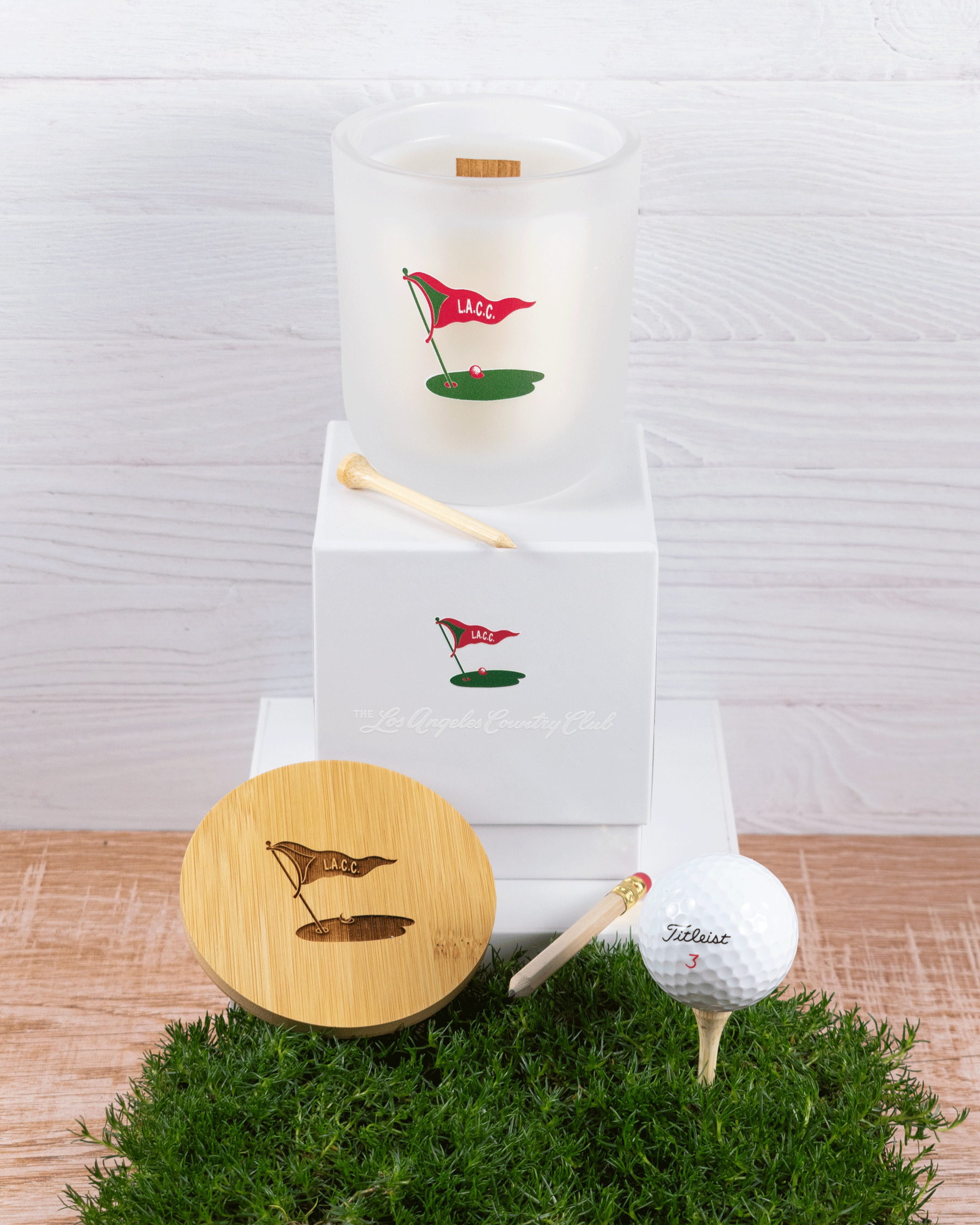 A 12 oz Luxe Candle in "Cashmere" a diffused white from Custom Candles L.A with white wax and a wooden wick. The candle is printed with the "L.A.C.C." logo in red & green of a golf flag, ball, and green. It sits next to a white tee on a white box with same logo and "Los Angeles Country Club printed. Leaning on the left side of the box is a wooden candle lid that is engraved with the same logo. They sit on a patch of grass next to a pencil and a teed up golf ball on a wooden floor and white wood background.