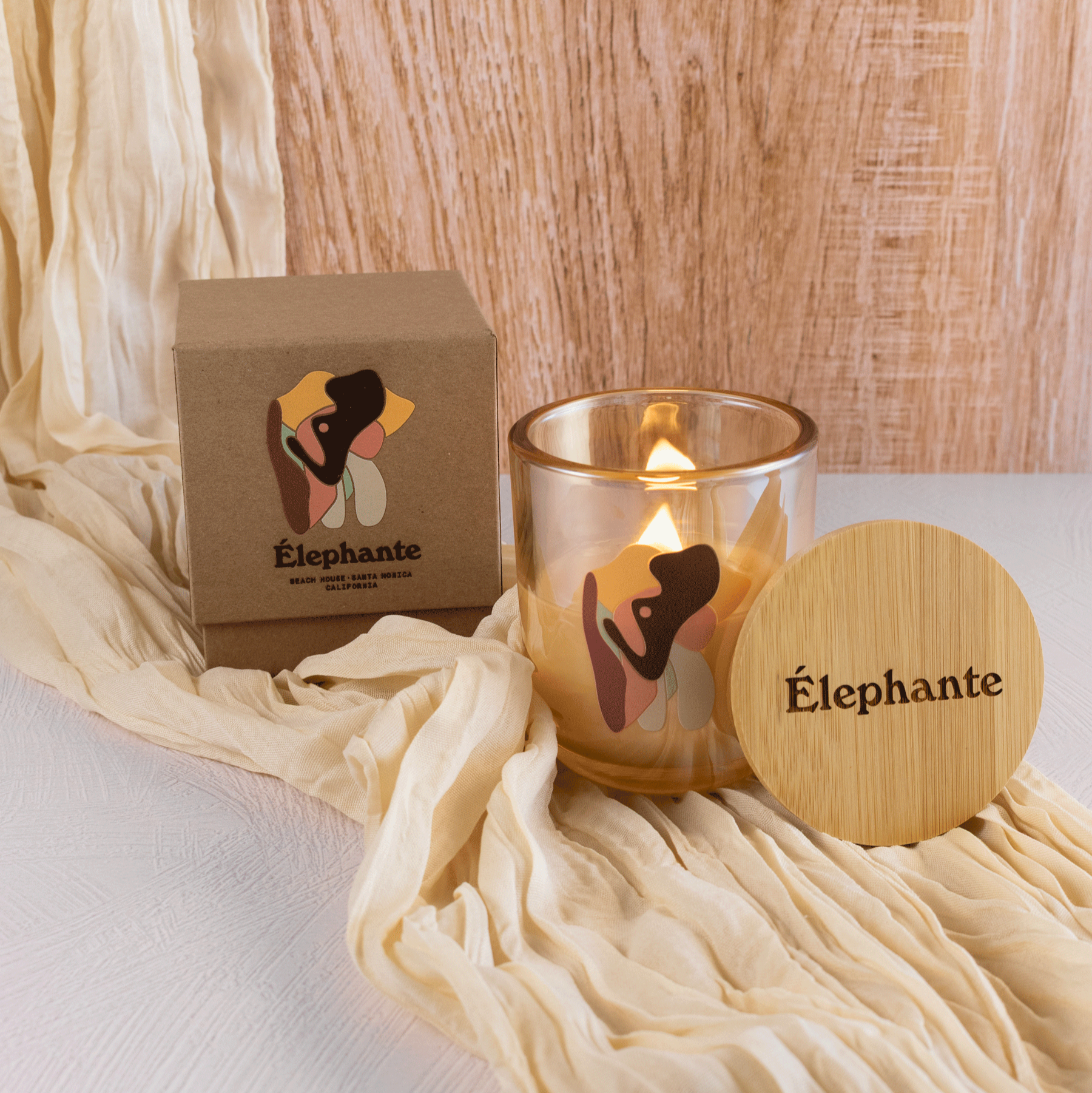 A lit 12 oz Luxe Candle in "Daybreak" a glimmering gold color from Custom Candles L.A. The candle is printed with a mosaic "Elephant" logo in yellow, brown, teal, pink, and grey. On the left sits a Kraft box printed with the same image and reads "Elephante" below the mosaic. On the right of the candle leans a wooden candle lid that is engraved with the word "Elephante". All three items sit on a cream colored table runner on top of a white textured ground, and a wooden background.
