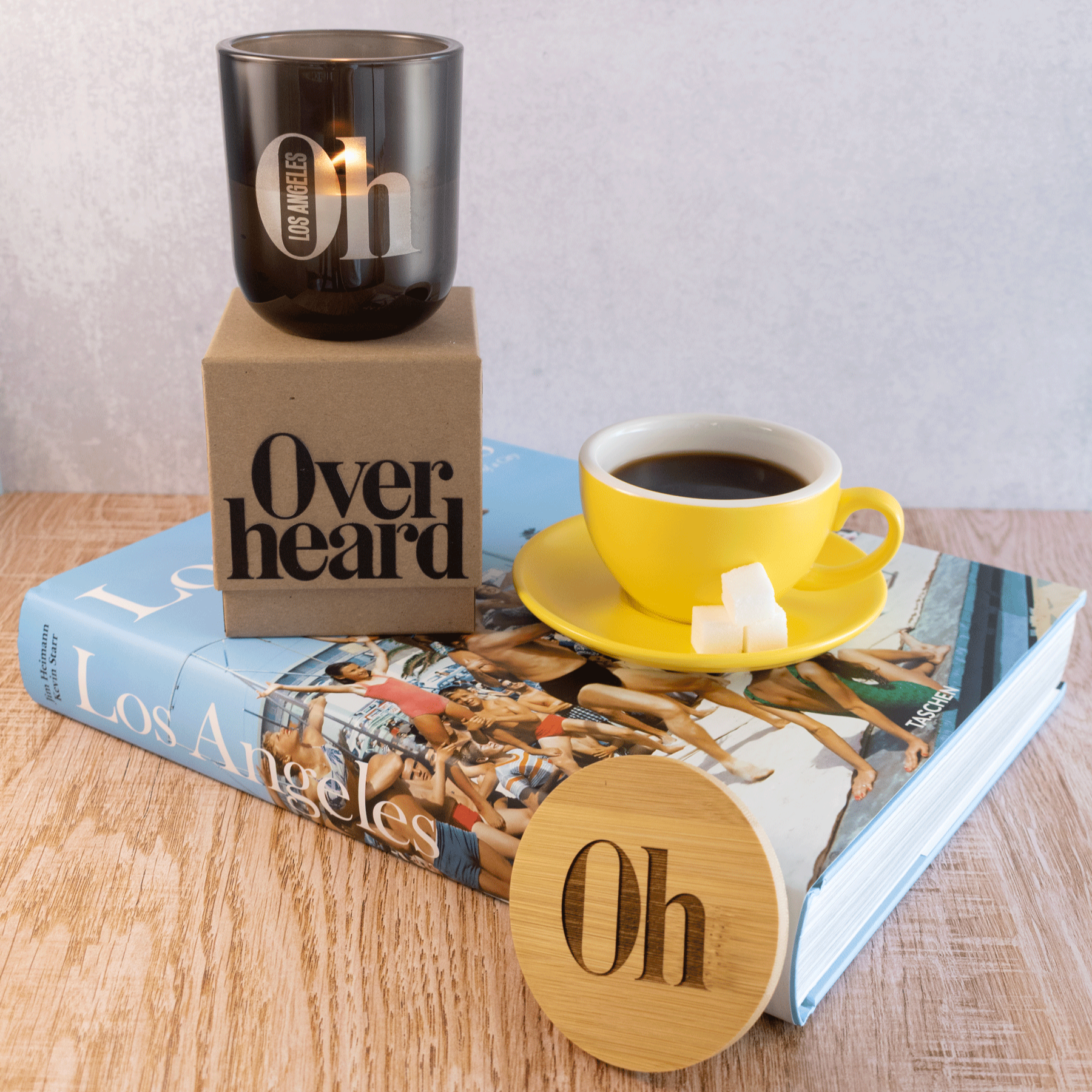 A 12 oz Luxe Candle in "Nightfall" a smoky grey color from Custom Candles L.A. The candle is etched with a logo that reads "Oh Los Angeles" with the flame shining through. It sits on top of a Kraft box with the logo that reads "Overheard" printed in black. They both sit on a thick book titled "Los Angeles" along with a cup of coffee in a yellow mug. Leaning against the book is a wooden lid to the candle that is engraved with "Oh". The book sits on a wooden table with a gray wall in the background.