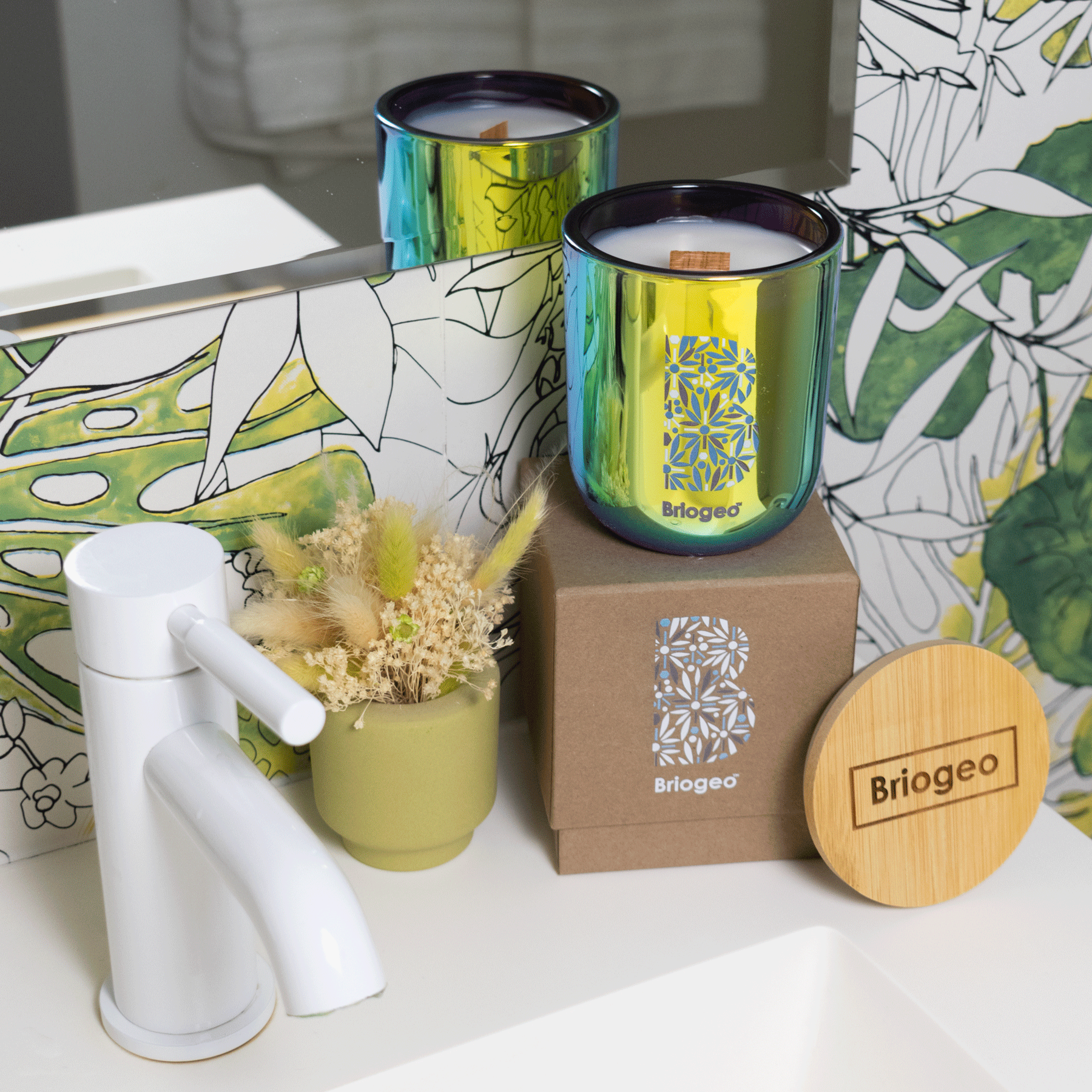 A 12 oz Luxe Candle in "Euphoria" a metallic blue color from Custom Candles L.A with white wax and a wooden wick. The candle is printed with the "Briogeo" logo in blue and grey. It sits on a kraft box printed with the same logo in grey, white, and blue. Leaning on the right side of the box is a wooden candle lid engraved with "Briogeo" inside a rectangle. The items all sit next to a plant on a white sink with a white faucet. The background is a wall with a plant-patterned wallpaper in green and a mirror.