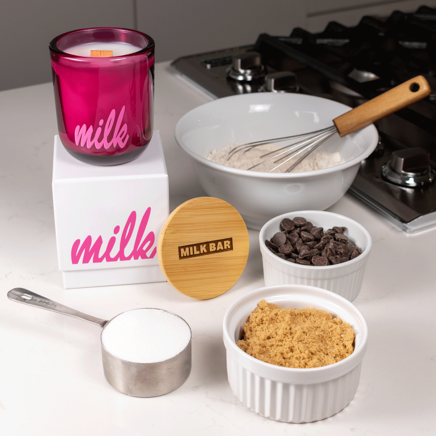 A 12 oz Luxe Candle in "Cherry Kiss" a juicy magenta color from Custom Candles L.A with white wax and a wooden wick. The candle is printed with a logo that reads "milk" in pink. It sits on a white box with the same logo printed in pink. Leaning on the right side of the box is a wooden candle lid that is engraved with a logo that reads "Milk Bar" in a shaded rectangle. They sit on a white countertop next to a stove, surrounded by a bowl of flour with a whisk, chocolate chips, brown sugar, and white sugar.