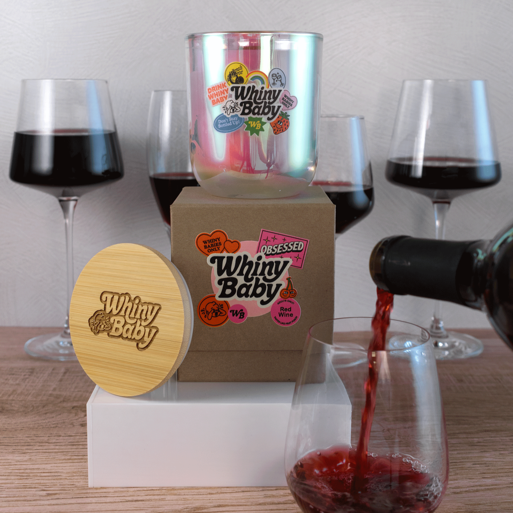 A 12 oz Luxe Candle in "Bliss" an iridescent pink from Custom Candles L.A. The candle is printed with a logo that reads "Whiny Baby" in black, with multicolored "Whiny Baby" stickers surrounding. It sits on a Kraft box with the Whiny Baby logo in various pinks, black, and white. Leaning on the left side of the box is a wooden candle lid that is engraved with the logo. All three items sit up on a white box. A bottle of red wine is being poured in the front, and there are 4 glasses of red wine behind.