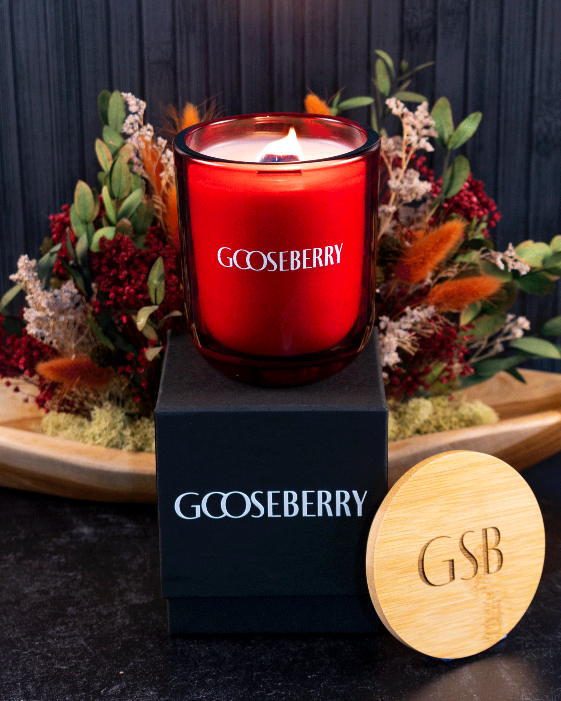 A lit 12 oz Luxe Candle in "Blood Red" a vivid rouge color from Custom Candles L.A with white wax and a wooden wick. The candle is printed with a logo that reads "Gooseberry" in white. It sits on a black box with the same logo printed in white. Leaning on the right side of the box is a wooden candle lid that is engraved with "GSB". All three items sit in front of a bouquet of red, green and orange stems on a black floor, in front of a black slat background.