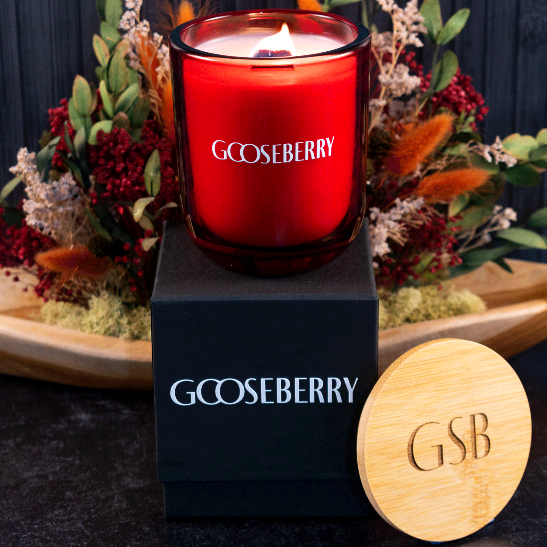 A lit 12 oz Luxe Candle in "Blood Red" a vivid rouge color from Custom Candles L.A with white wax and a wooden wick. The candle is printed with a logo that reads "Gooseberry" in white. It sits on a black box with the same logo printed in white. Leaning on the right side of the box is a wooden candle lid that is engraved with "GSB". All three items sit in front of a bouquet of red, green and orange stems on a black floor, in front of a black slat background.