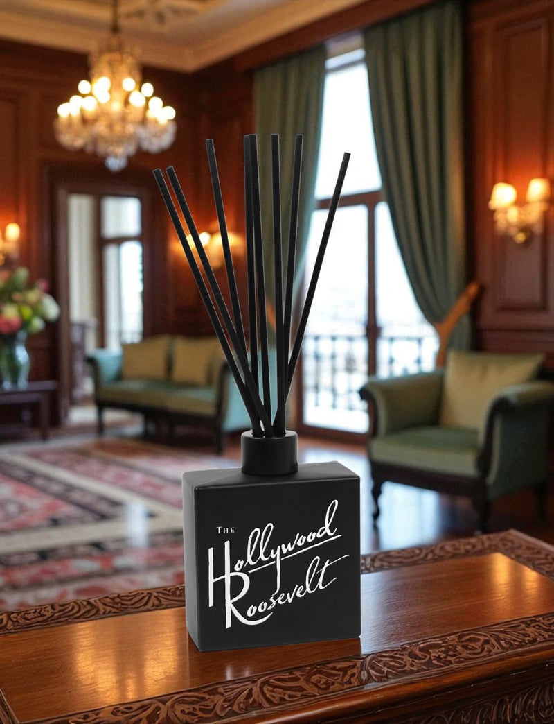 A black reed diffuser with white script text and black reed sticks sits on an ornate carved wooden coffee table in an elegant hotel lobby, with crystal chandeliers, rich wood paneling, green velvet curtains, and classic furnishings in the background