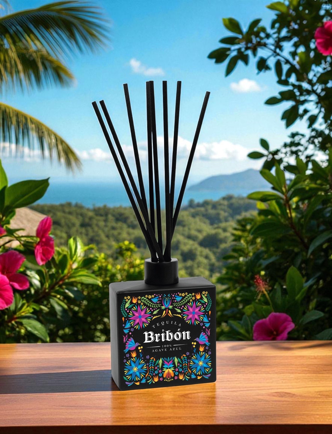 A black reed diffuser with colorful floral pattern branding sits on a wooden deck table, surrounded by tropical foliage including palm fronds and pink hibiscus flowers, with lush green mountains and blue sky in the background