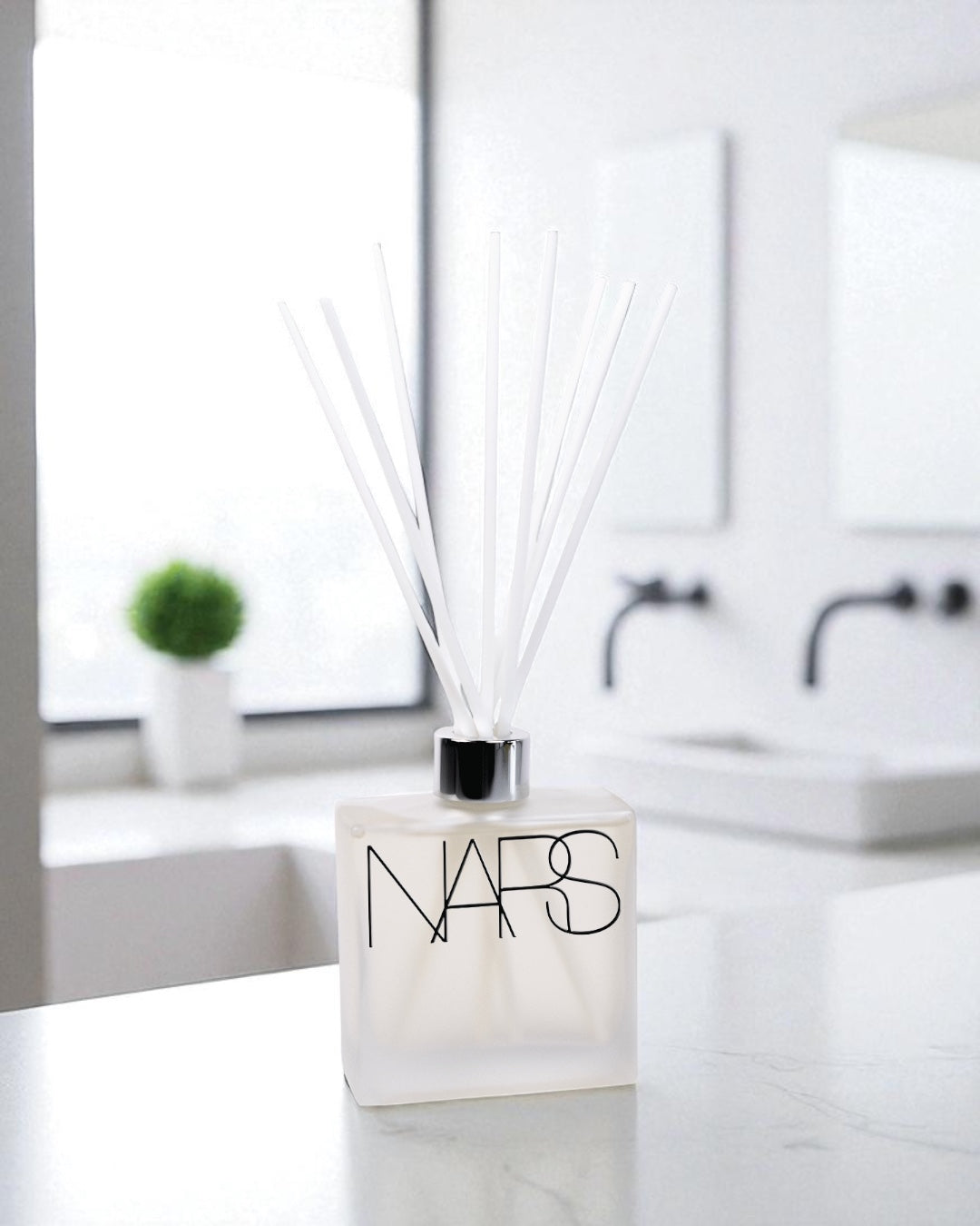 A frosted glass reed diffuser with black text and white reed sticks sits on a white marble countertop in a modern bathroom, with white subway tile backsplash, black fixtures, and a small green plant visible in the background