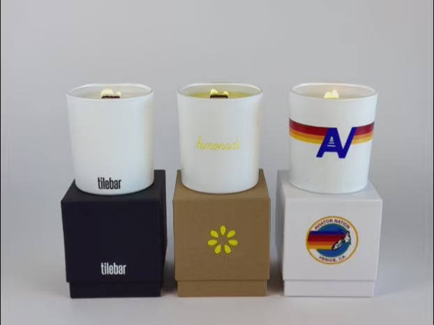 Load video: video showcase of candle lighting of 3 white 14 oz candles on top of their respective gift box, all in a row