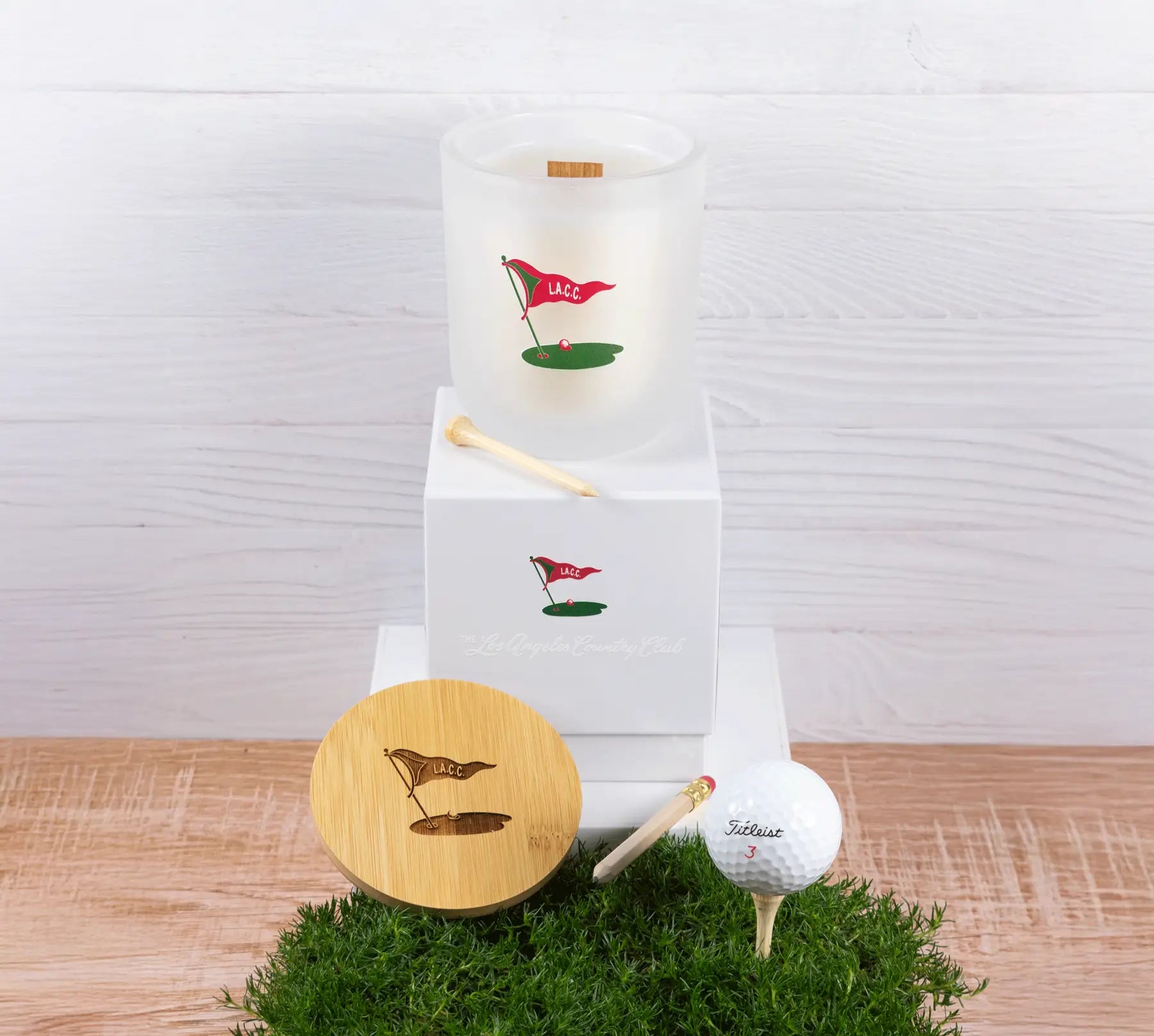 Golf-themed candle, golf ball, and club on a grassy surface with a white background