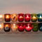 A display of colorful glass candles, some lit and some unlit, arranged in two rows on clear shelves against a plain wall. Each candle has a unique design or text on it.