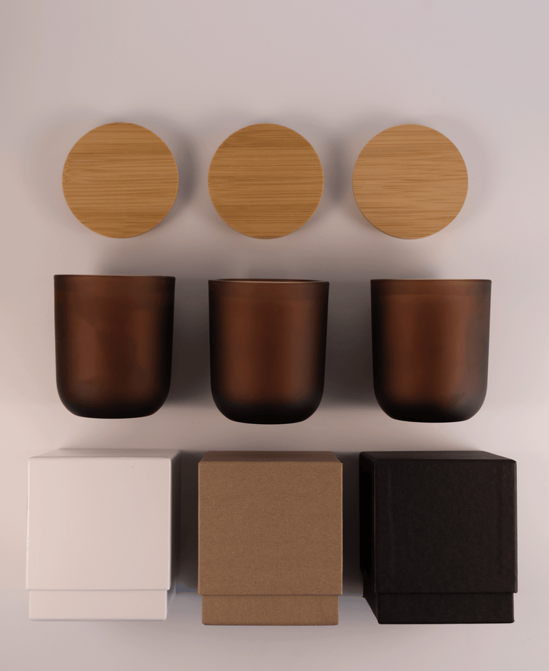 Three amber glass tumblers with wooden lids above and three different boxes; white, kraft, and black; on a white surface.