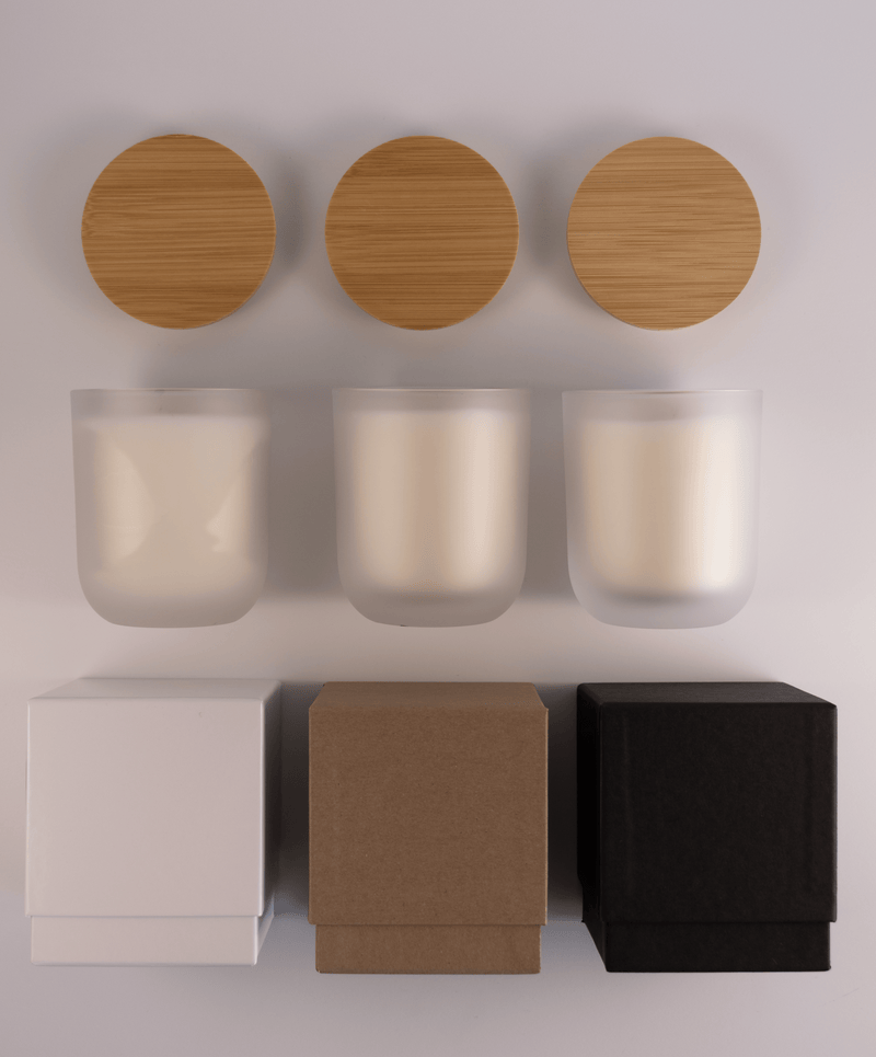 Three frosted glass tumblers with wooden lids above and three different boxes; white, kraft, and black; on a white surface.