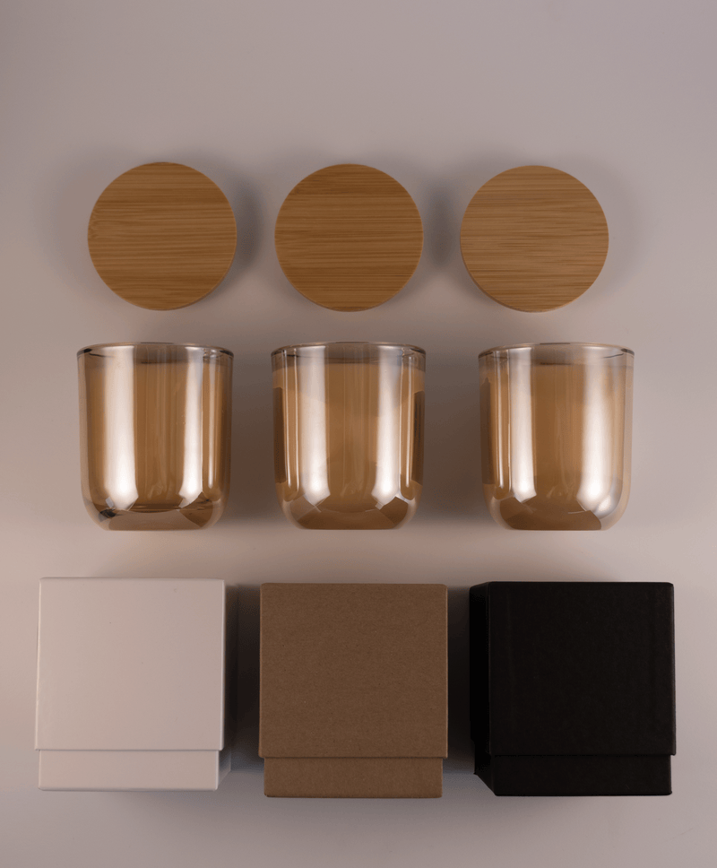 Three champagne gold glass tumblers with wooden lids above and three different boxes; white, kraft, and black; on a white surface.