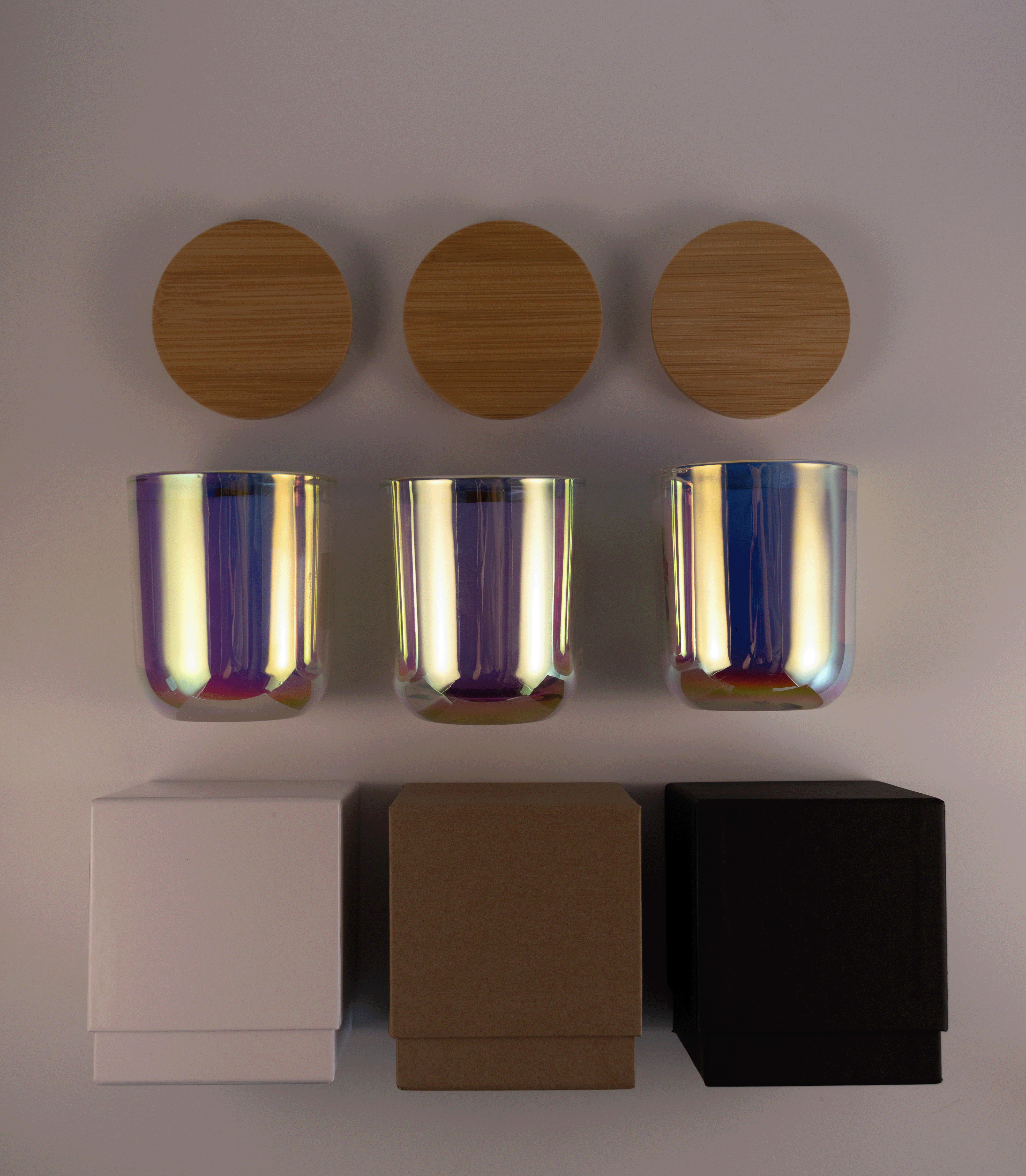 Three iridescent pink glass tumblers with wooden lids above and three different boxes; white, kraft, and black; on a white surface.
