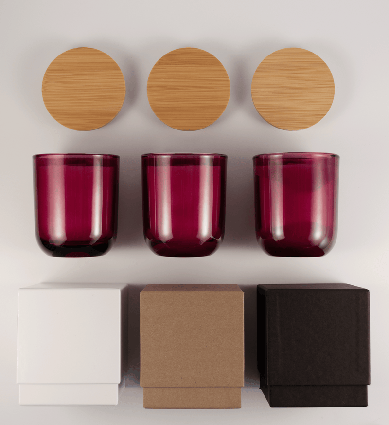 Three magenta glass tumblers with wooden lids above and three different boxes; white, kraft, and black; on a white surface.