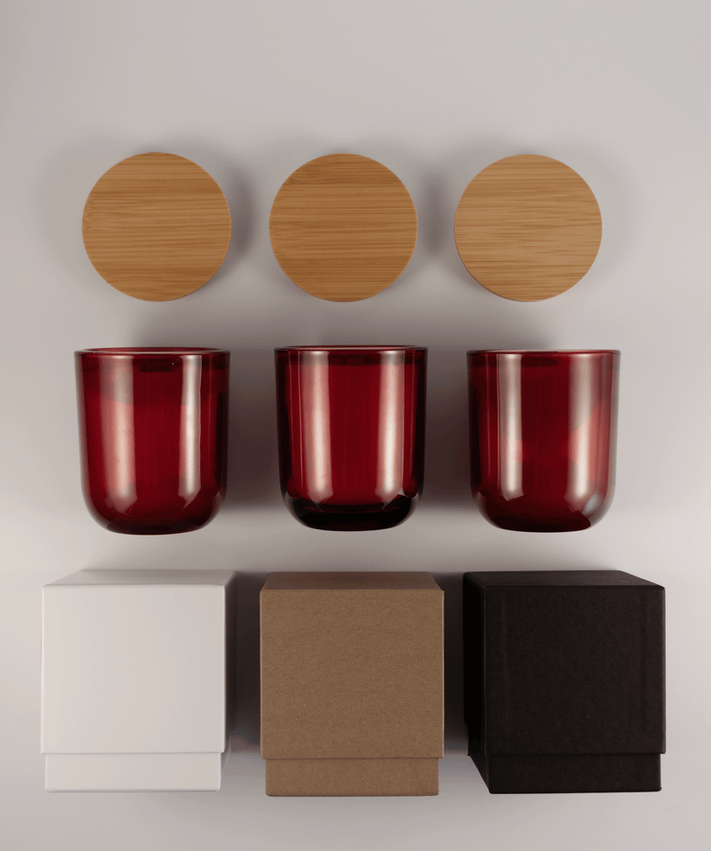 Three red glass tumblers with wooden lids above and three different boxes; white, kraft, and black; on a white surface.