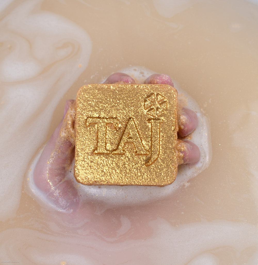 Hand holding a gold square bath bomb with a debossed logo in foamy bath water