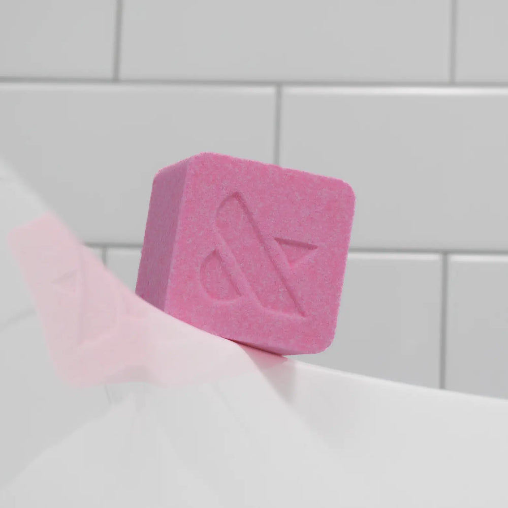 Pink square bath bomb with a debossed logo resting on the edge of a bathtub