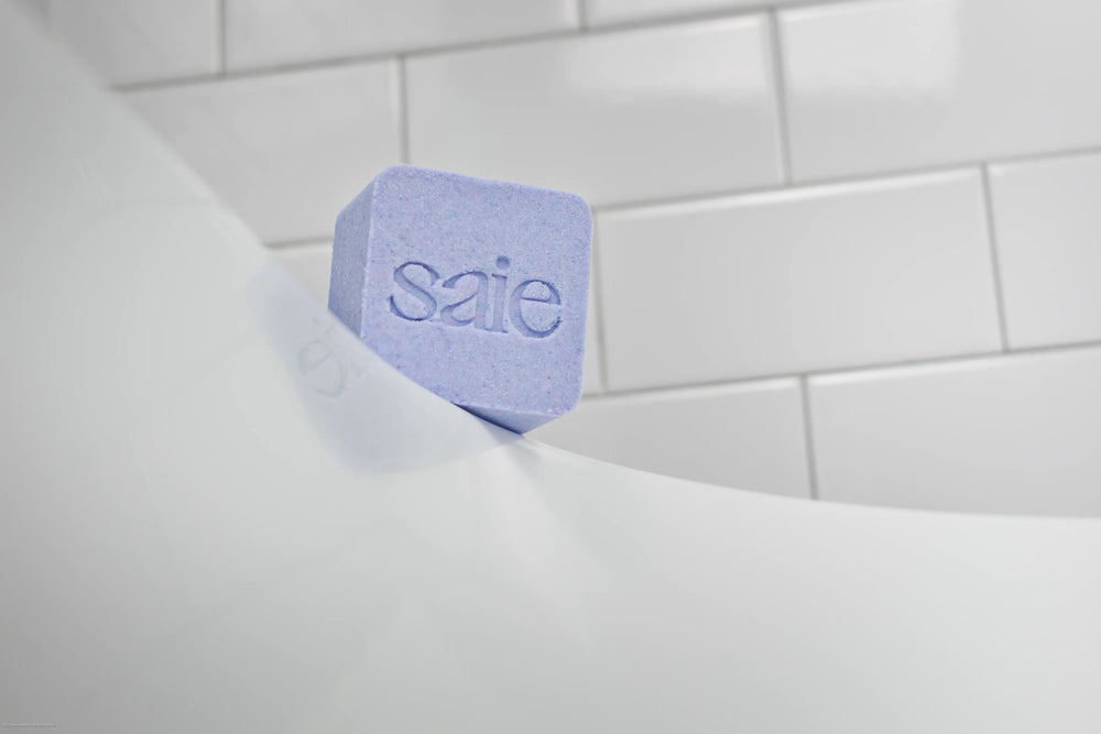Purple square bath bomb with a debossed logo resting on the edge of a bathtub