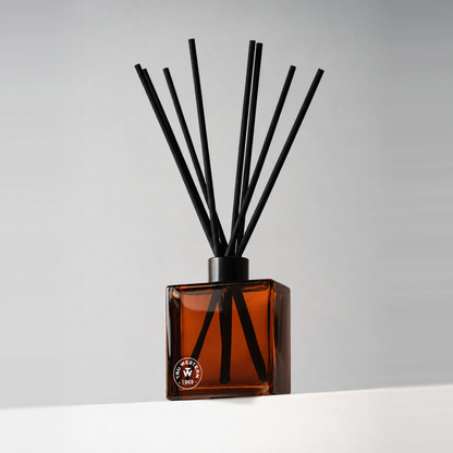Dark tinted glass 8 oz. reed diffuser with black reeds and a small printed logo, on a white ledge against a soft gray backdrop