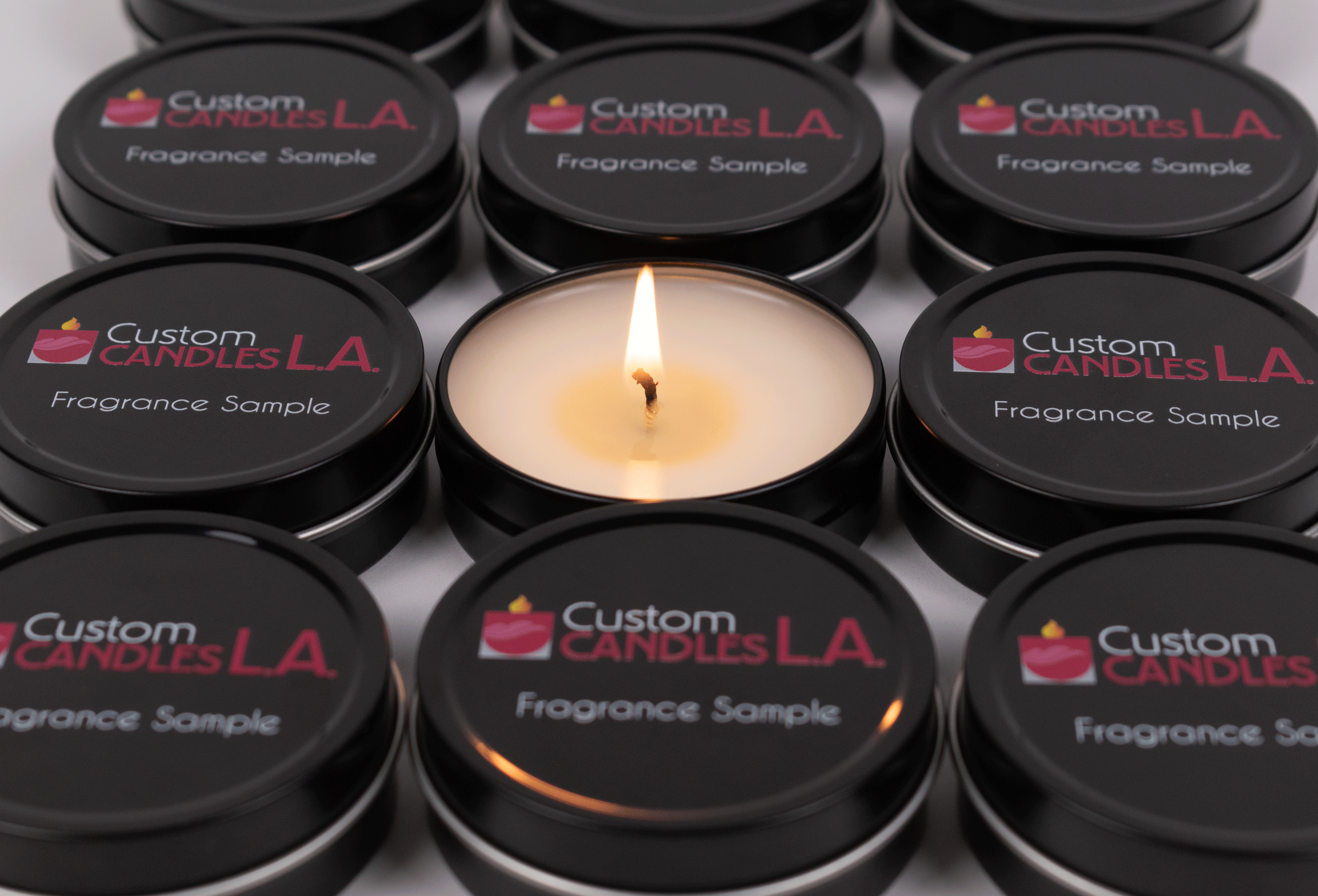 A 3/4 angle close up view of 12 black fragrance sample tins from Custom Candles L.A. aligned neatly in rows of three. Each tin has the Custom Candles L.A. logo printed on it in white and burgundy with the "Fragrance Sample" printed in white below the logo. The fragrance sample in the middle has its lid removed and is lit, showing a small flame and the white wax, partially melted, and a slightly burned wick.
