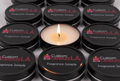 A 3/4 angle close up view of 12 black fragrance sample tins from Custom Candles L.A. aligned neatly in rows of three. Each tin has the Custom Candles L.A. logo printed on it in white and burgundy with the "Fragrance Sample" printed in white below the logo. The fragrance sample in the middle has its lid removed and is lit, showing a small flame and the white wax, partially melted, and a slightly burned wick.