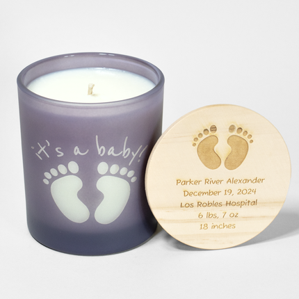 The image shows a 14 oz frosted lavender candle from Custom Candles LA. The candle features baby footprints and the text "it's a baby!" where the white wax shows through the glass. Next to the candle is a wooden lid engraved with baby footprints and the text that states the baby's name, birthday, birth place, weight and height. The candle is unlit, with a single wick visible, making it a charming gift for celebrating a baby's arrival.