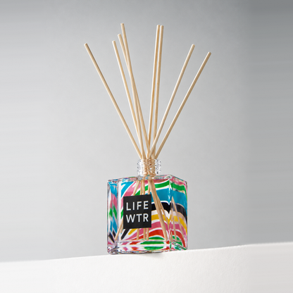 Clear glass 8 oz. reed diffuser with natural reeds and a full-color printed wrap, on a white ledge against a soft gray backdrop