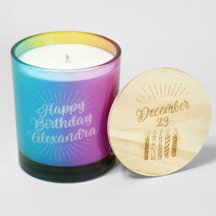 A rainbow colored 14 oz. candle from Custom Candles LA with "Happy Birthday Alexandra" etched on the glass, coupled with a wooden lid engraved with "December 23" and candle illustrations.