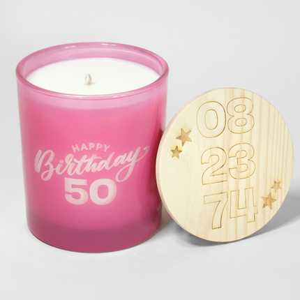 A pink premium Happy Birthday Candle with Personalized Age—Premium in a glass holder with "Happy Birthday 50" written on it. The lid, placed on its side, is wooden and features carved numbers "08," "23," and "74" alongside star shapes. This Custom Candles LA custom candle makes for a perfect personalized age gift. The background is plain and white.