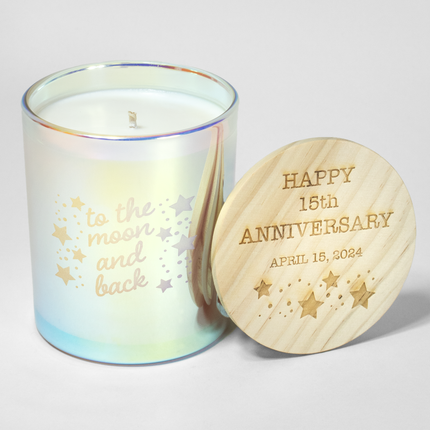 The "To the Moon and Back Anniversary" candle is featured in an iridescent candle from Custom Candles LA, with the text "to the moon and back" etched into the glass with stars and dots surrounding the text. Next to it sits a wooden lid that is engraved with the text "Happy 15th Anniversary April 15, 2024" with stars and dots below the text.