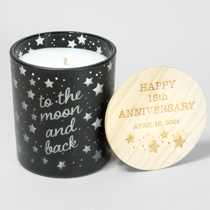 A 14 oz black vessel candle from Custom Candles LA. The holder features etched stars on the glass and the phrase "to the moon and back" with white wax showing through.  Next to the candle is a wooden lid engraved with "HAPPY 15th ANNIVERSARY APRIL 15, 2024" and star motifs. The unlit candle has a single visible wick, making it a romantic and celebratory anniversary gift.