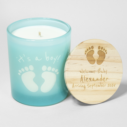 The image shows a 14 oz Malibu blue candle from Custom Candles LA. The candle features baby footprints and the text "it's a boy!" where the white wax shows through the glass. Next to the candle is a wooden lid engraved with baby footprints and the message "Welcome Baby Alexander Arriving September 2025." The candle is unlit, with a single wick visible, making it a charming gift for celebrating a baby boy's arrival.