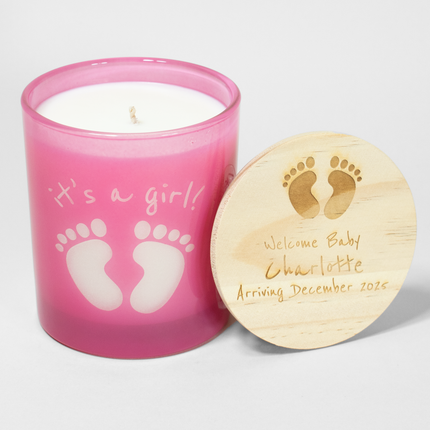 The image shows a 14 oz pink candle from Custom Candles LA. The candle features baby footprints and the text "it's a girl!" where the white wax shows through the glass. Next to the candle is a wooden lid engraved with baby footprints and the message "Welcome Baby Charlotte Arriving December 2025." The candle is unlit, with a single wick visible, making it a charming gift for celebrating a baby girl’s arrival.