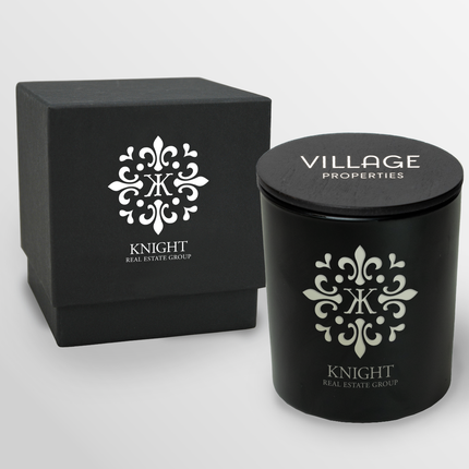 A 14 oz Deluxe "Design Your Own" Candle in a black glass vessel from Custom Candles LA. The candle features an etched logo that reads "Knight Real Estate Group" on the glass vessel and a black wooden lid imprinted with white text that reads "Village Properties".  Behind the candle is a 2 piece black box with a logo that reads "Knight Real Estate Group" printed in white. 