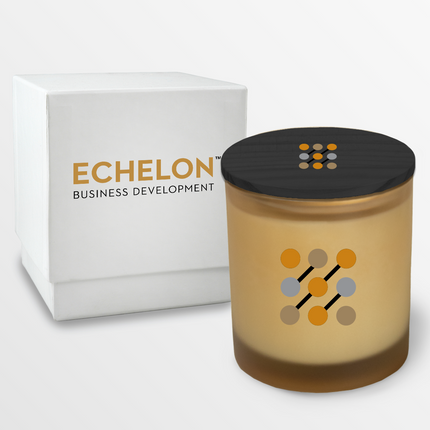 A 14 oz Deluxe "Design Your Own" Candle in a toasted coconut glass vessel from Custom Candles LA. The candle features a 4 color imprinted logo on the glass vessel and black wooden lid. Behind the candle is a 2 piece white box with an imprinted logo that reads "Echelon Business Development"