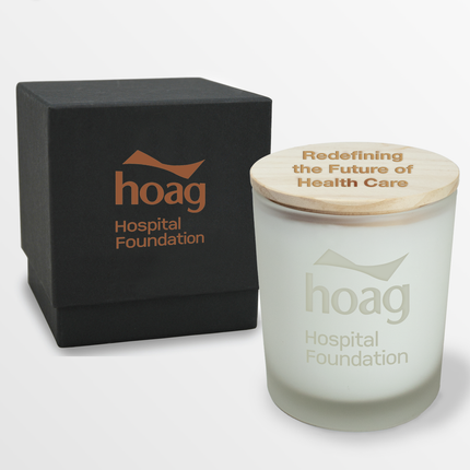 A 14 oz Deluxe "Design Your Own" Candle in an frosted white glass vessel from Custom Candles LA. The candle features an etched logo reading "hoag Hospital Foundation" on the glass vessel and a wooden lid imprinted with the text "Redefining the Future of Health Care" in burnt orange. Behind the candle is a 2 piece black box with a the logo "Hoag Hospital Foundation" in burnt orange.